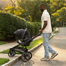 Nuna - Triv Lx + Pipa Urbn travel system in caviar pushed by a man along a sidewalk in warm lighting.