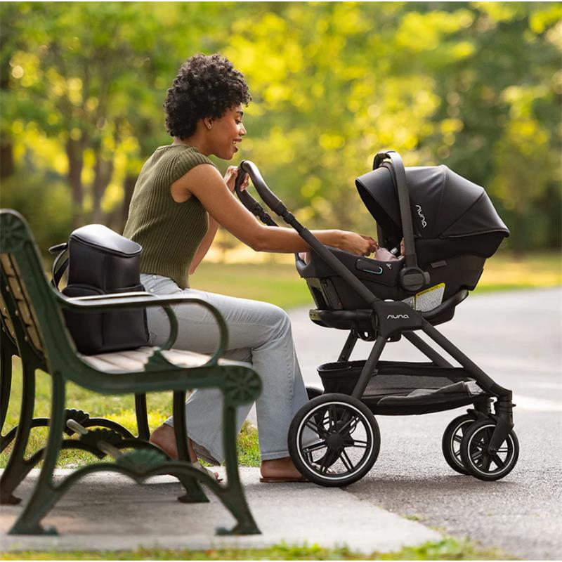 A woman interacts with a Nuna Triv Lx stroller featuring the Pipa Urbn infant seat, outdoors on a sunny day.