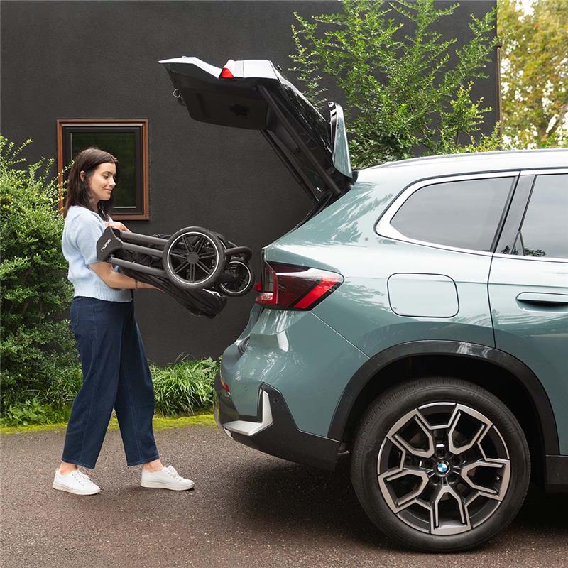 A woman places a folded Nuna Triv Lx stroller into the trunk of a BMW SUV parked outdoors.