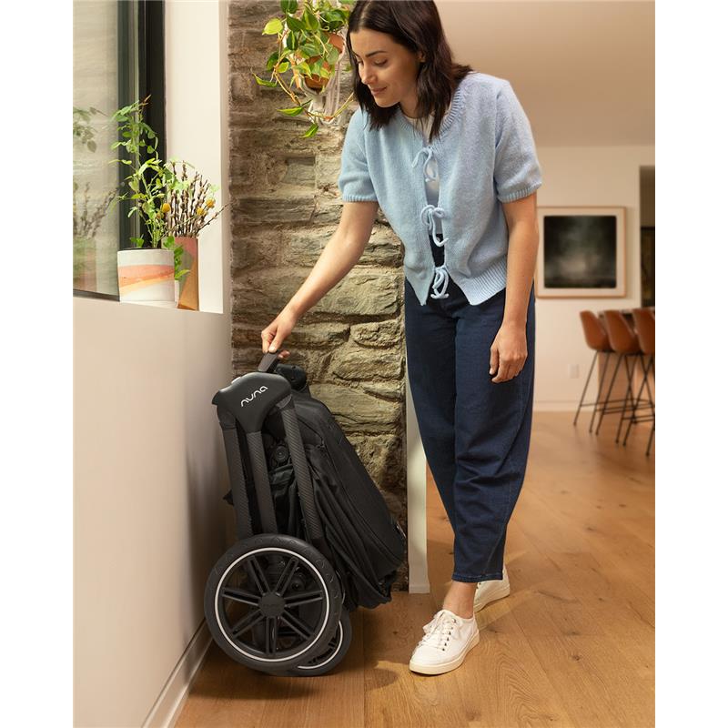 Nuna Triv Lx stroller in caviar color, folded and leaning against a wall, with a woman adjusting it in a modern indoor setting.