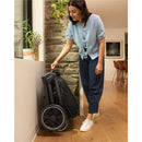 Nuna Triv Lx stroller in caviar color, folded and leaning against a wall, with a woman adjusting it in a modern indoor setting.
