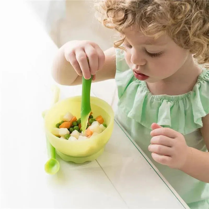 A child is using a green soft-tip training spoon to eat from a bowl of mixed vegetables.