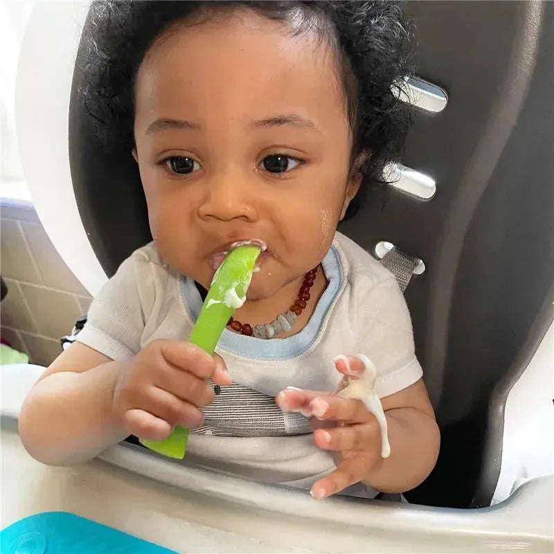 A baby with curly hair is sitting in a high chair, using a green silicone training spoon while eating.
