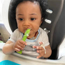 A baby with curly hair is sitting in a high chair, using a green silicone training spoon while eating.