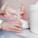 Hand holding the Ola Baby diaper rash cream applicator, with a suction base, coral color, near a changing pad and storage basket.