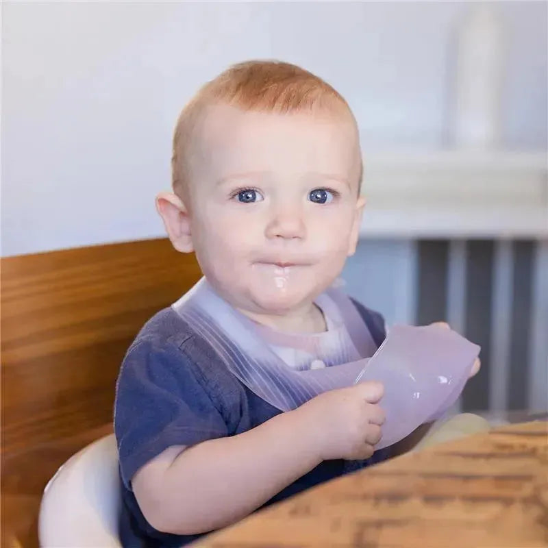Baby wearing a lilac silicone waterproof bib, seated at a table, holding a bib with a slight smile.