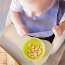 Silicone baby bib in use, with a baby seated in a high chair, holding a green bowl of cereal.