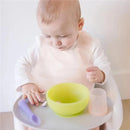 A baby wearing a lilac waterproof bib sits in a high chair, playing with a green bowl, spoon, and a cup.