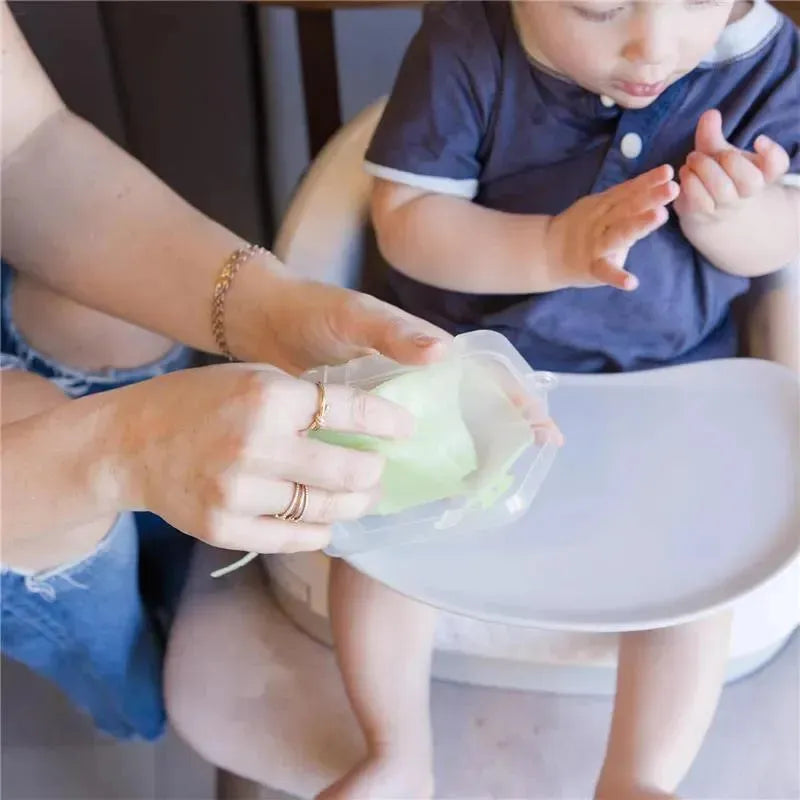 A parent holds a lilac-foldable silicone baby bib while a toddler plays on a high chair.