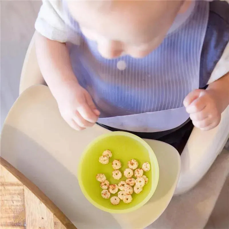 Silicone baby bib in lilac color worn by a child, sitting at a high chair with a bowl of snacks.