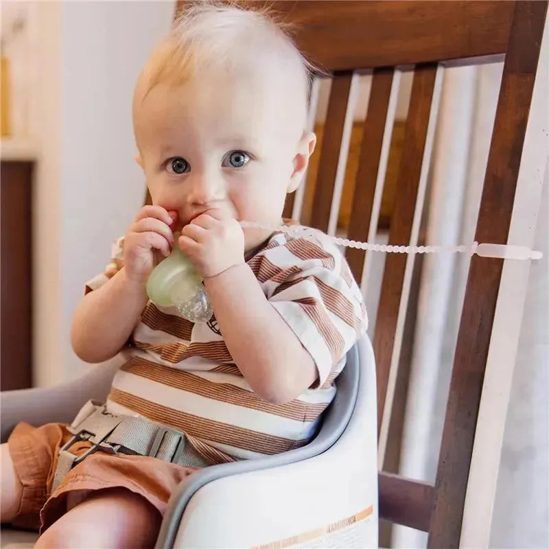 Baby in a high chair holding a silicone strap connected to a green container, wearing a striped shirt and looking curiously.