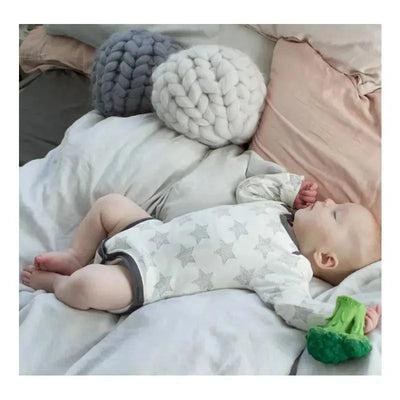Infant lying on a bed, holding a green chewable toy shaped like broccoli while surrounded by soft blankets.