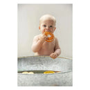 A baby in a metal tub holds and bites a chewable orange toy, Clementino The Orange by Oli & Carol.