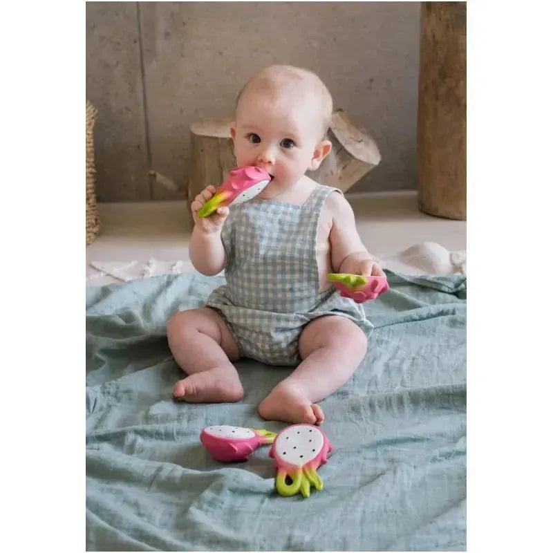 A baby sits on a light blue blanket, chewing on a fuchsia dragon fruit chewable toy, with two additional toys beside them.