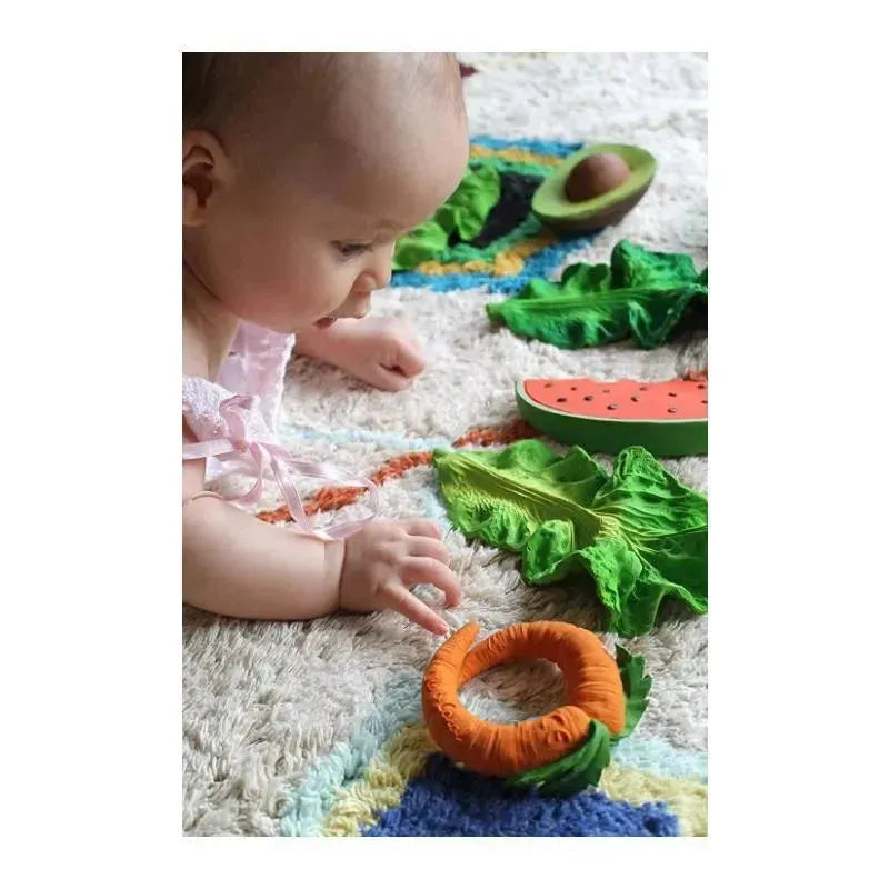 Infant reaching for the Oli & Carol chewable toy, Kendall The Kale, amidst vibrant vegetable-themed toys on a textured rug.