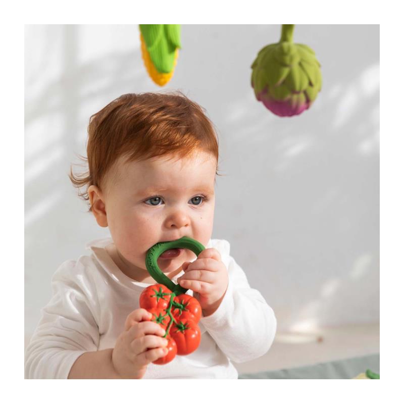 A baby with red hair bites a green-handled rattle toy shaped like a cluster of red tomatoes.