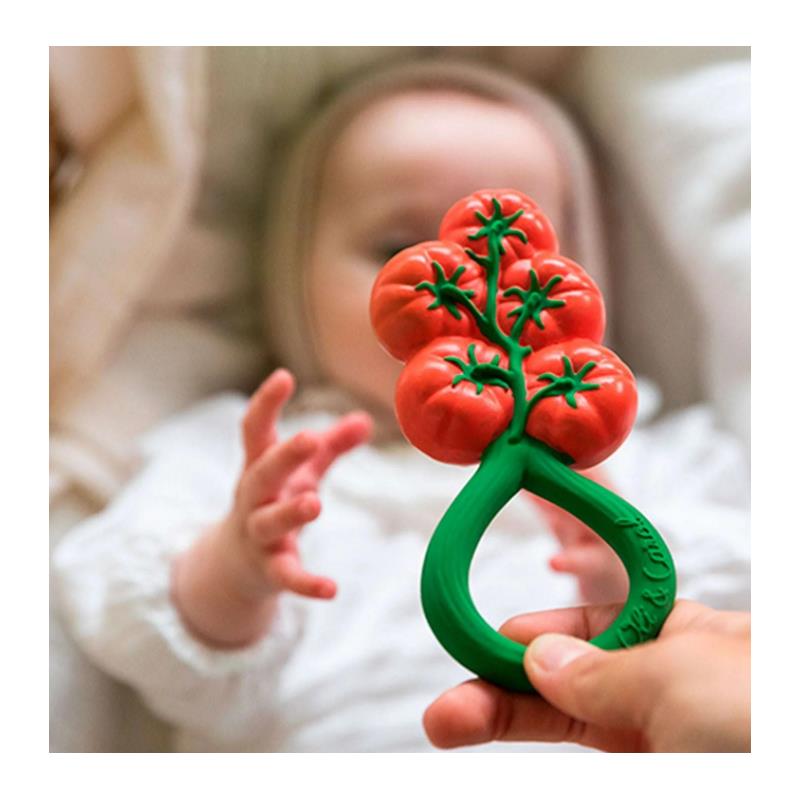 Oli & Carol tomato-shaped rattle toy held in a hand, with a baby reaching out in the background.