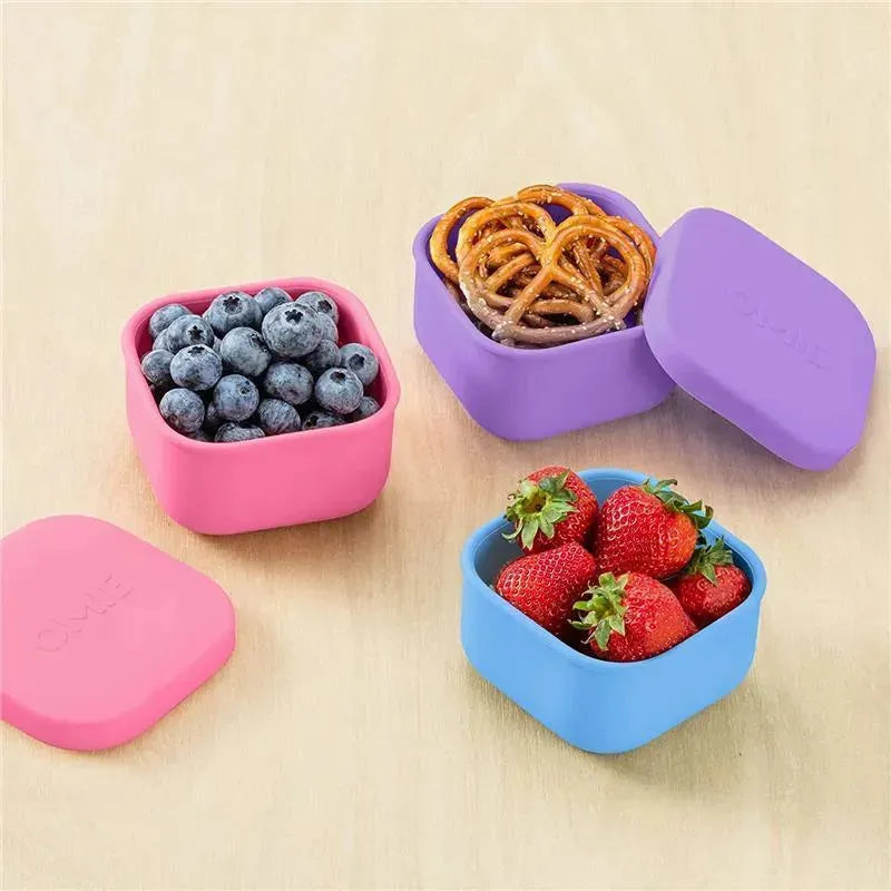 Purple OmieBox food storage container with lid, filled with pretzels, surrounded by containers of blueberries and strawberries.