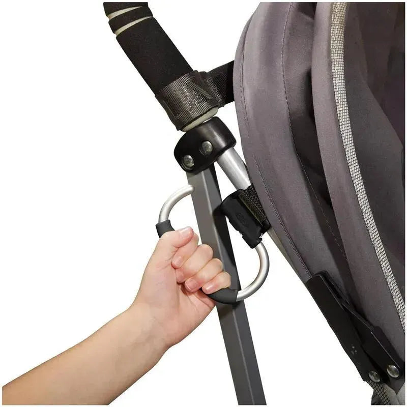 Hand gripping an Oxo grey stroller hook attached to a stroller frame, demonstrating its functionality for carrying items.