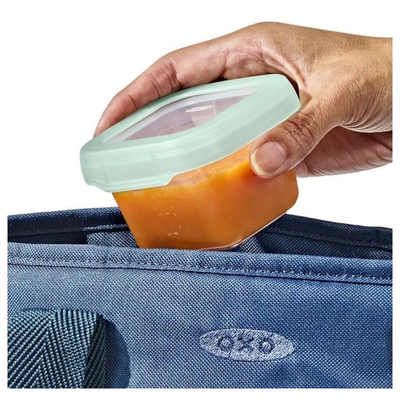Hand holding an Oxo Tot food storage container filled with orange baby food, placing it into a blue bag.