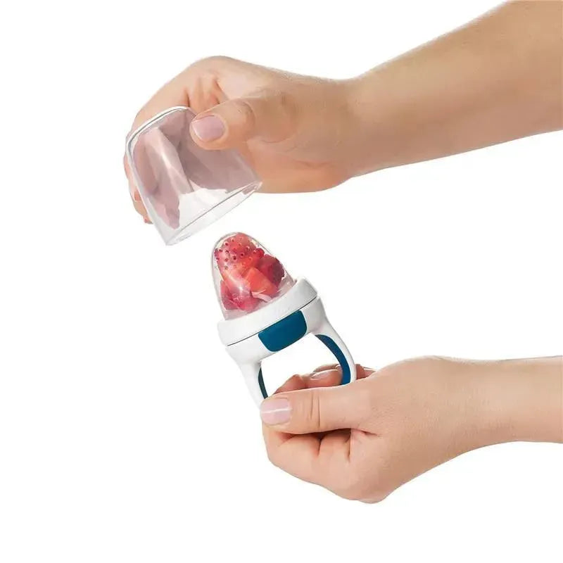 Two hands holding an OXO silicone feeder replacement, with a clear cap removed, revealing pieces of fruit inside.