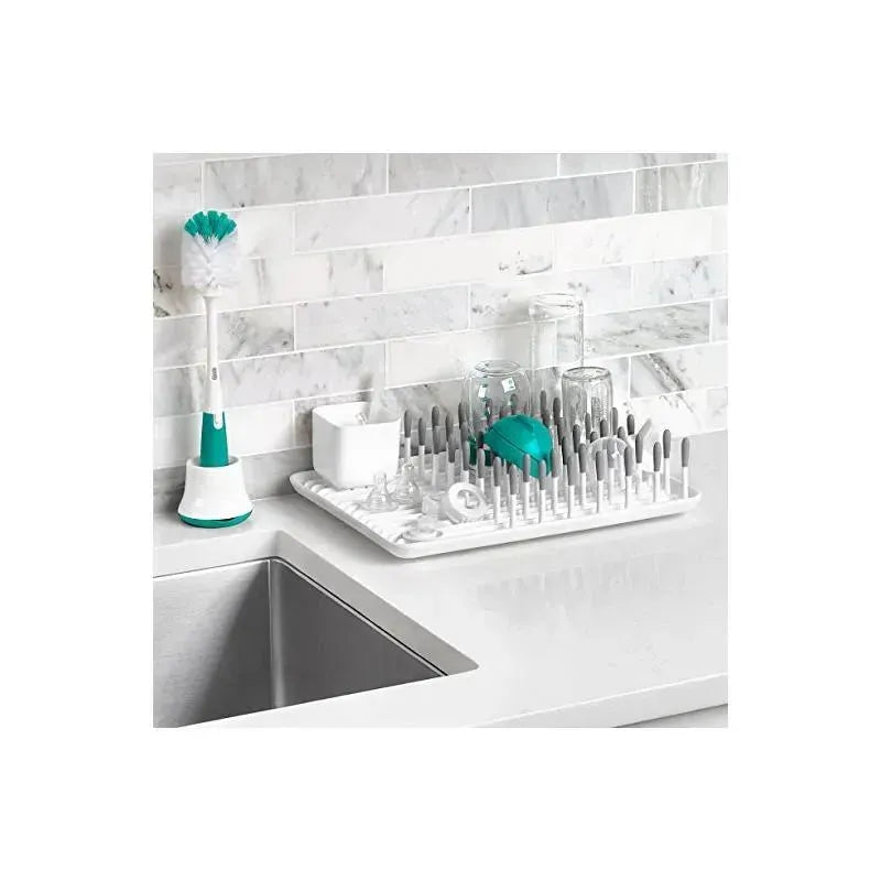 OXO Tot Bottle Drying Rack in gray with multiple prongs, bottles, and a bottle brush on a kitchen countertop.