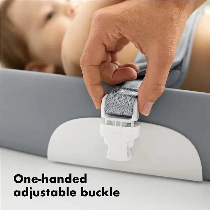 Hand adjusting the one-handed adjustable buckle on an OXO Tot Changing Pad, with a baby in the background.