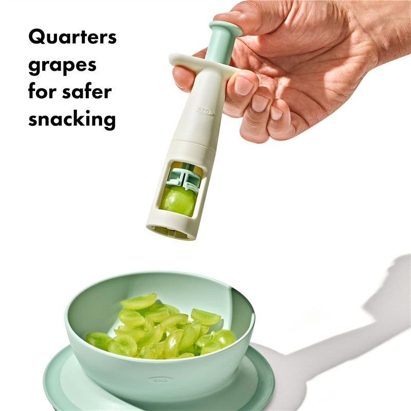 OXO Tot Grape Cutter in opal color; quarters grapes for safer snacking, held above a bowl with sliced grapes.