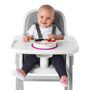A grinning baby sits in a high chair, with a pink Oxo Tot Stick & Stay Divided Plate filled with fruit and cereal in front.