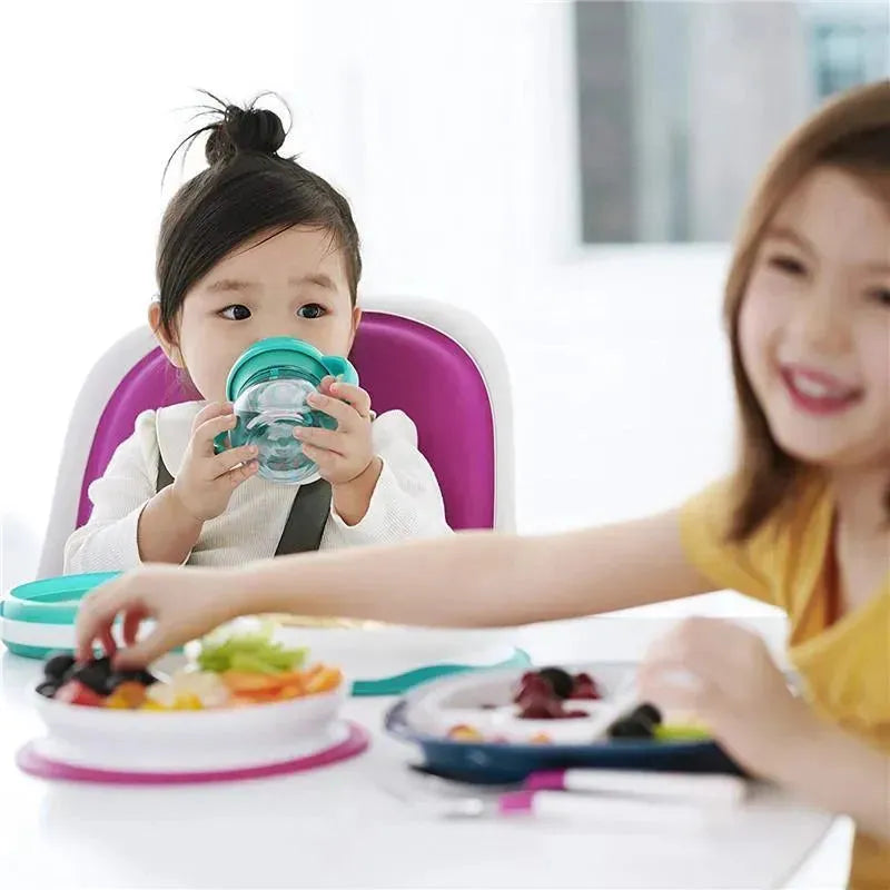 Divided pink plate from Oxo - Tot, with kids enjoying meals; one child sips from a cup while reaching for food.