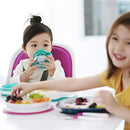 Divided pink plate from Oxo - Tot, with kids enjoying meals; one child sips from a cup while reaching for food.