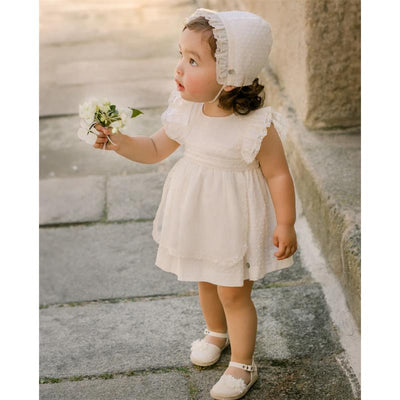 Baby girl wearing a cream woven dress with ruffled sleeves, holding a small bouquet of flowers in a scenic outdoor setting.