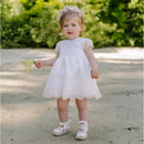 Toddler wearing a cream dress with lace trim and a floral headband, holding a small bouquet of flowers.