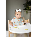 Infant wearing a woodland-themed bib sits in a high chair, holding food, with a neutral background and plants behind.