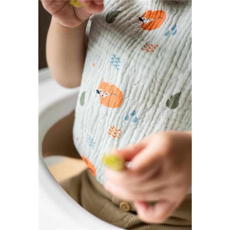 Baby wearing a muslin cotton bib featuring woodland animals, specifically a fox design, while holding a snack.
