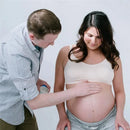 A woman in a light tank top smiles as a man applies a molding agent to her pregnant belly, demonstrating the Pearhead Belly Casting Kit.