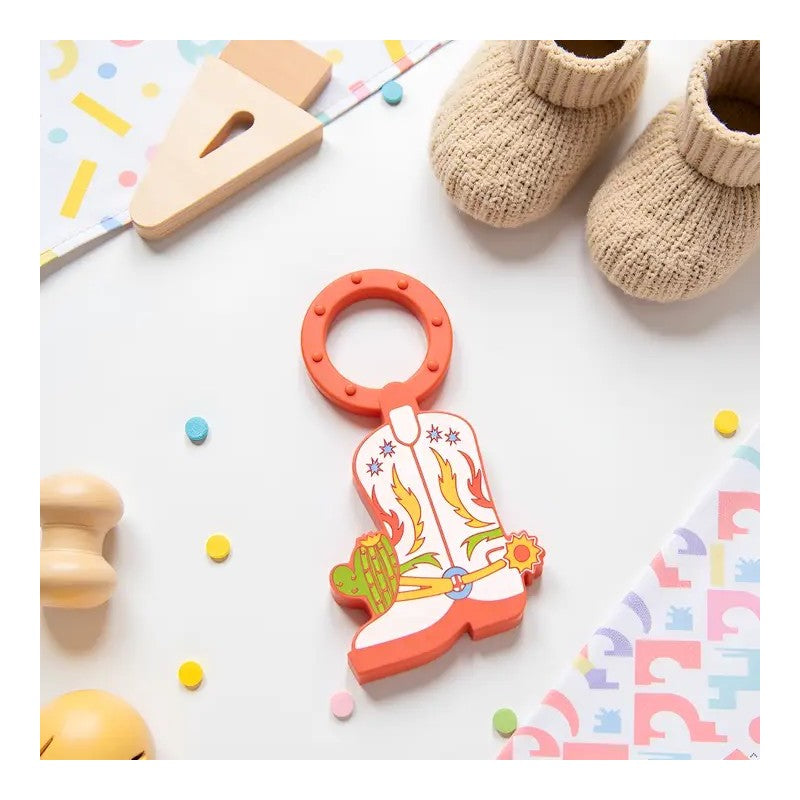 Cowboy boot-shaped teether ring in orange and white with decorative colors, surrounded by baby items on a playful backdrop.
