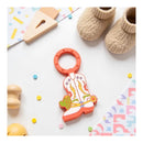 Cowboy boot-shaped teether ring in orange and white with decorative colors, surrounded by baby items on a playful backdrop.