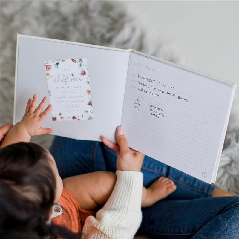 Ivory linen baby memory book opened to a page with a baby shower invitation and a checklist, held by a caregiver and child.