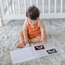 Baby in an orange romper, sitting on a fluffy rug, examining the Pearhead Ivory Linen Baby Memory Book open to photos and notes.