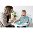 Mother feeding a smiling baby in a high chair, both wearing a silicone bib from the Pearhead Milk Drunk set.