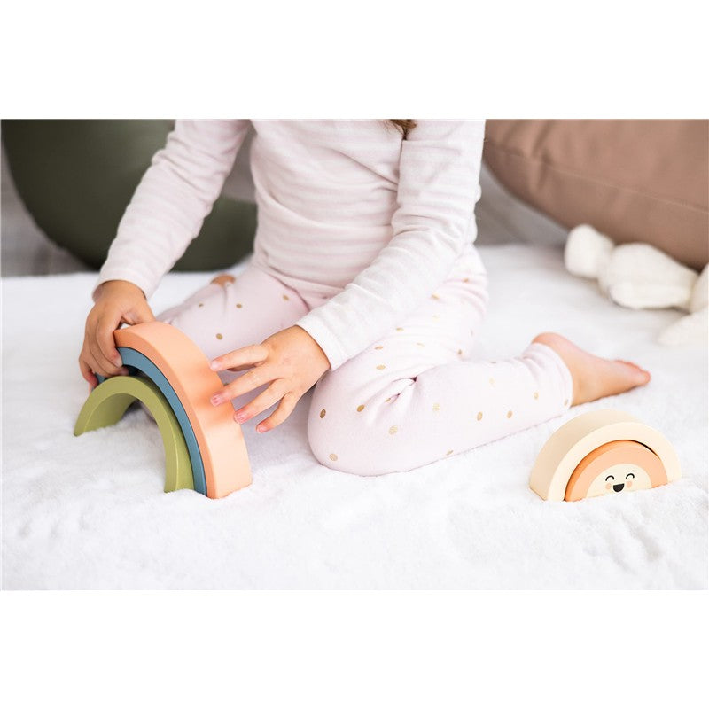 A child in pajamas builds with the Pearhead Wooden Rainbow Stacking Toy, featuring colorful arch shapes on a soft surface.