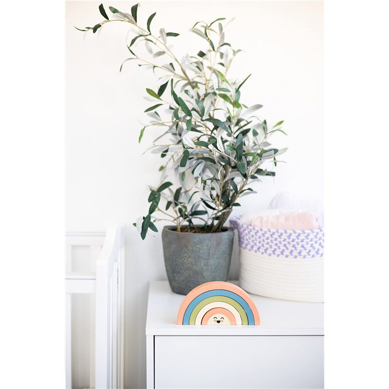 Wooden rainbow stacking toy featuring pastel colors and a smiling face, placed beside a green potted plant.