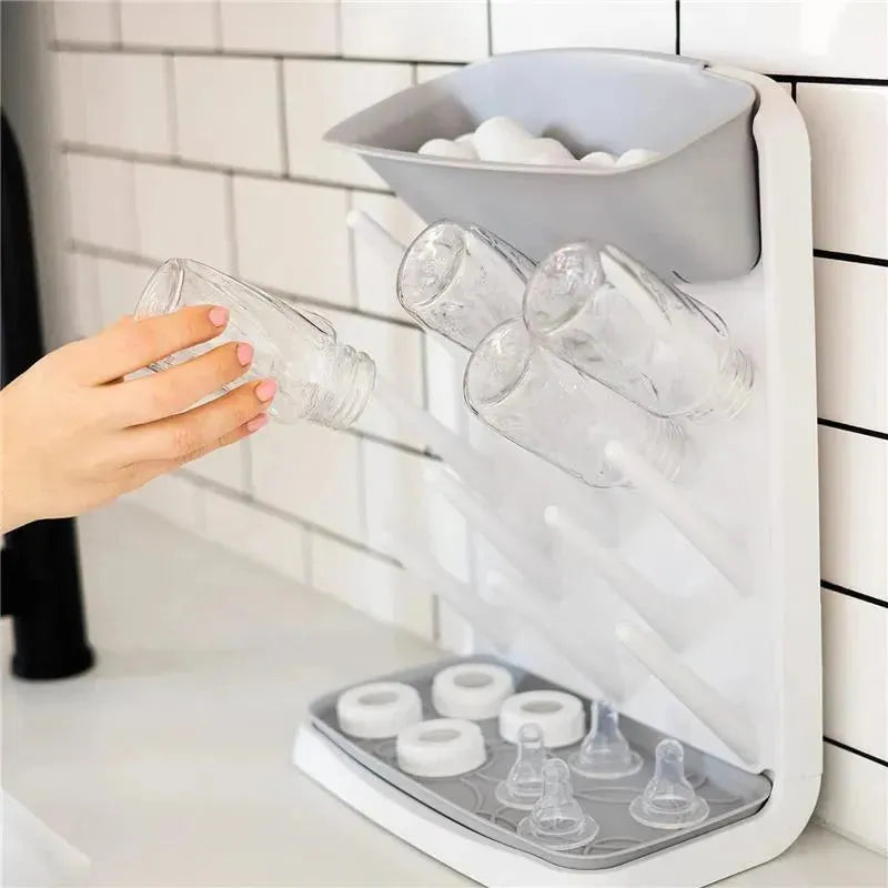Gray Ubbi Vertical Baby Bottle Drying Rack displaying clear bottles, a hand placing one, and slots for bottle parts.
