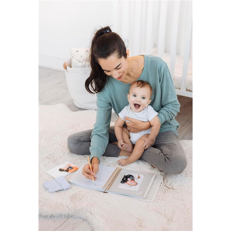 Mother in a teal sweater sits on a soft rug, writing in a Pearhead Deer Print Square Baby Book, holding a smiling baby.