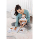 Mother in a teal sweater sits on a soft rug, writing in a Pearhead Deer Print Square Baby Book, holding a smiling baby.