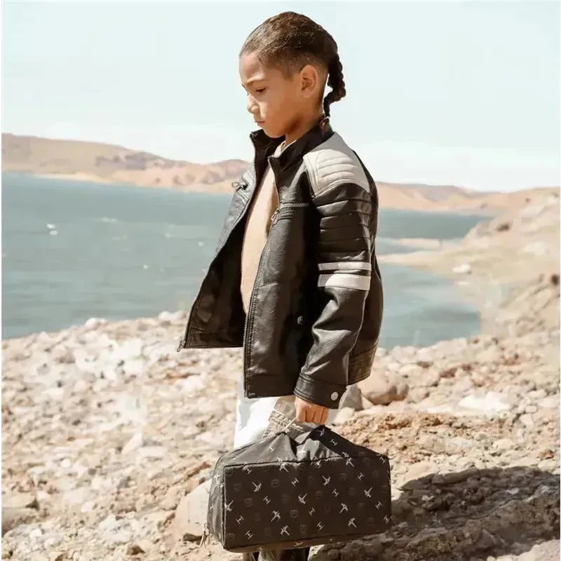 Young child wearing a black leather jacket stands on rocky terrain by the water, holding a patterned black bag.
