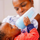 Baby drinks from a blue Pop Yum bottle, held by a child in red and white stripes, while an adult watches attentively.