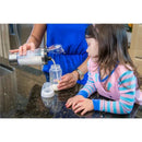A woman pours liquid from a stainless steel and acrylic mixer cup into a baby bottle while a child watches curiously.
