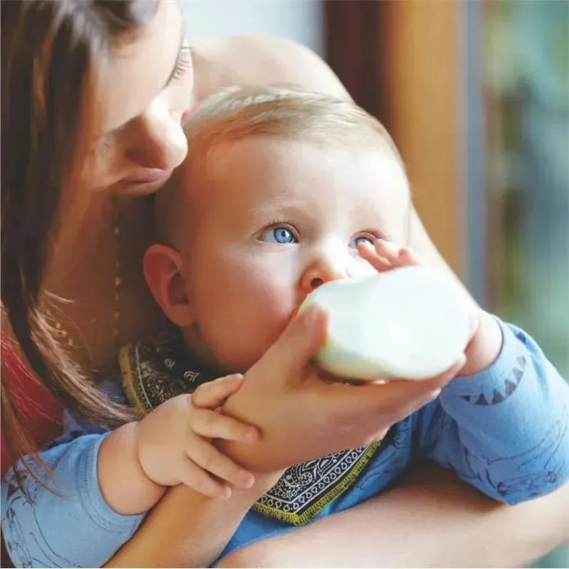 A parent feeds a baby with bright blue eyes from a formula mixer cup while sitting indoors.